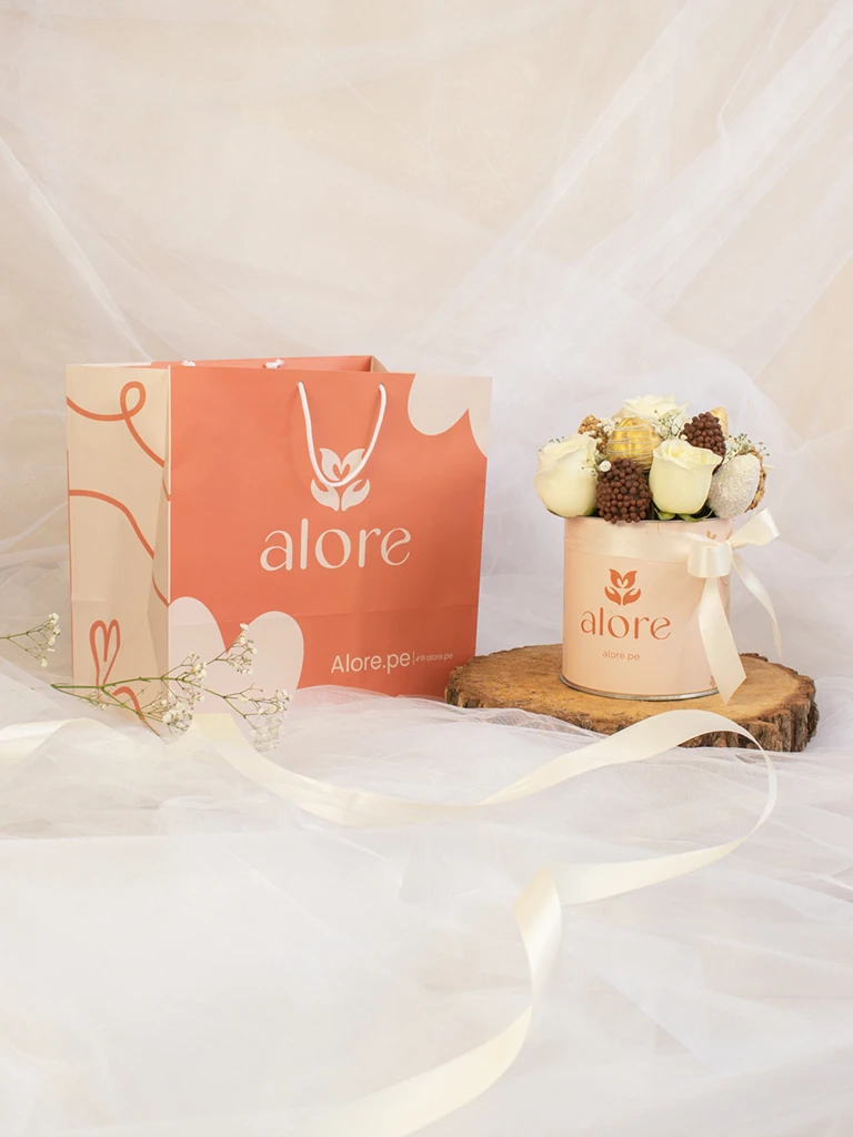 Caja pequeña de rosas blancas y fresas bañadas en chocolate sobre tronco junto a bolsa Alore