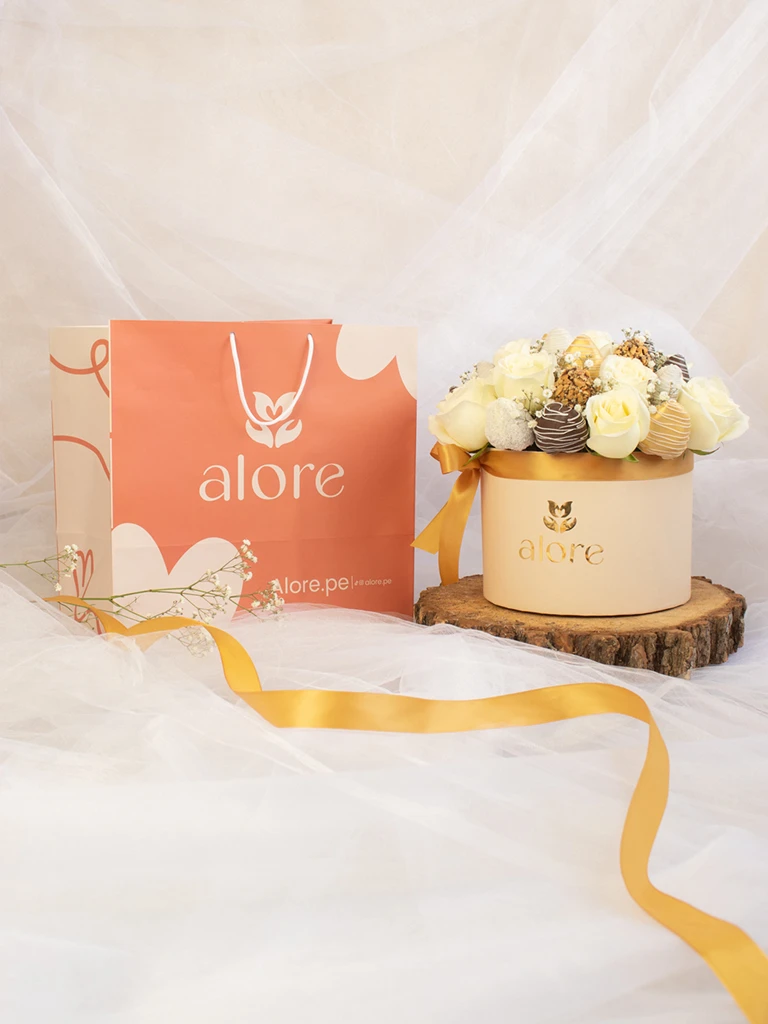 Caja grande de rosas blancas y fresas bañadas en chocolate con cinta dorada junto a bolsa Alore